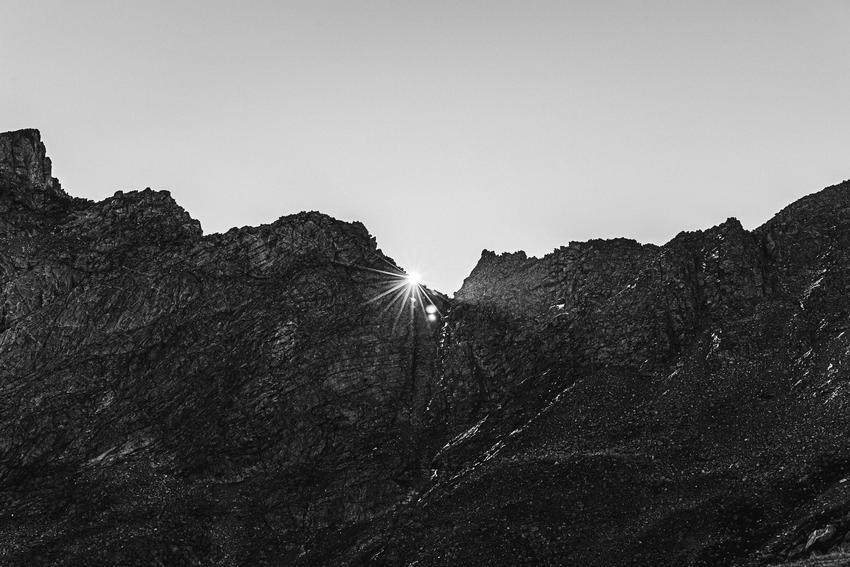 A black & white image of a sun shining through a crack of a mountain ridge