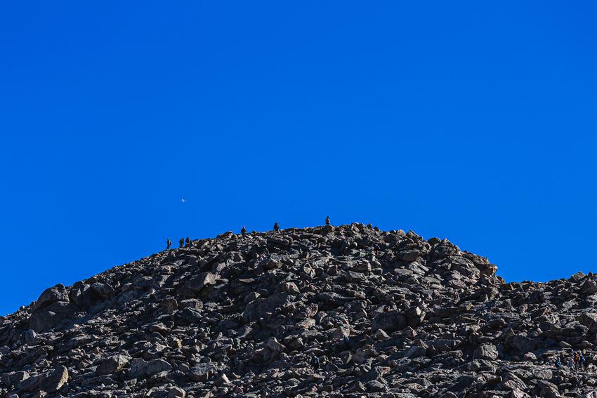 Rocky mountain summit with hikers on top. An airliner can be seen flying above